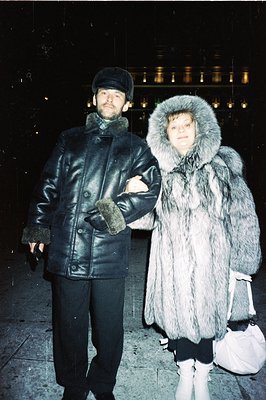 Man and woman pose outdoors, likely in a city setting based on the blurred background lights. Man wears a leather coat, cap, ...