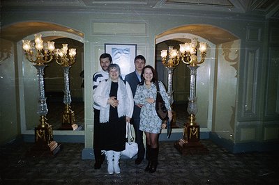 A group of four poses indoors, flanked by ornate, gilded sconces in an arched hall. The women wear 1990s-era fashion includin...
