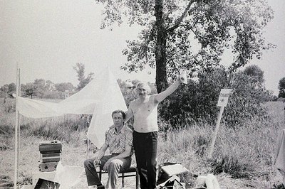 Two men outdoors; one seated, wearing a patterned shirt, the other standing shirtless with an arm raised. A simple fabric she...