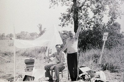A staged outdoor scene shows two men: one seated with documents, the other shirtless, raising a device overhead. Likely a fie...