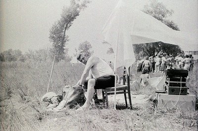 A man sits on a chair in tall grass, likely a film set, based on the background activity. A large cloth or backdrop is positi...