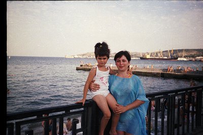 A young girl in a swimsuit and a woman in a blue dress stand on a seaside promenade. A crowd of bathers is visible on a nearb...