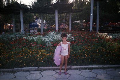 Young girl in a white swimsuit stands proudly in a vibrant flowerbed. A large pink inflatable ring is slung over her shoulder...