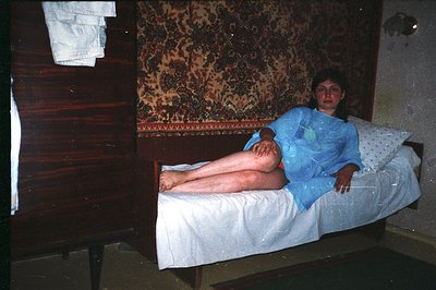 A woman lies on a vintage bed beneath an ornate, patterned rug, wearing a light blue medical gown. The room features dark woo...