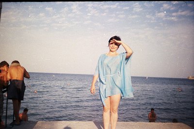 A woman stands on a concrete pier, facing the camera, shielding her eyes from the sun. She wears a short, light blue, tie-wai...