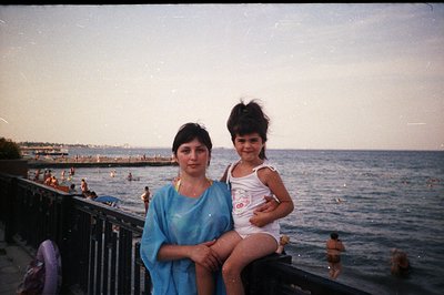 A woman carries a young girl on a seaside promenade. The scene captures a hazy sunset over the water, with blurred figures sw...