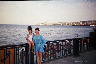 A woman and young girl pose by a seaside promenade, likely in Bulgaria. The woman wears a flowing, translucent blue dress; th...