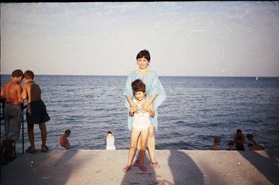 A young girl in a white dress and purple sandals stands being held by a woman in a light blue dress on a concrete promenade o...