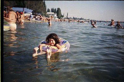 A young child floats on a colorful inflatable ring in a crowded seaside scene. Lush greenery and buildings line the backgroun...