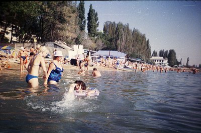 A bustling seaside scene, likely Bulgaria, captures a crowded beach with families enjoying the water. Sunbathers line the sho...