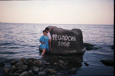 A woman and a child stand in shallow water beside a large, dark rock inscribed with Cyrillic text "Феодосия 2006". The rock s...