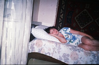 A person lies prone on a floral-patterned bed, eyes closed, wearing a blue and white patterned robe. The scene captures a dom...
