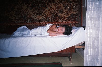 A young person reclines on a bed covered with crisp white linen, a richly patterned Oriental rug serving as the headboard. Th...