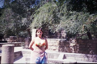 A woman stands outdoors, partially obscured by a large tree with green foliage, in a stone courtyard. She wears a floral-patt...