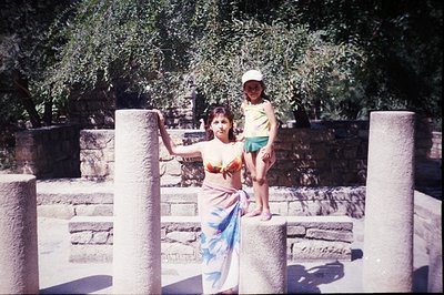 A woman and young girl pose amidst stone ruins, likely a seaside archeological site. The woman wears a patterned sarong and b...