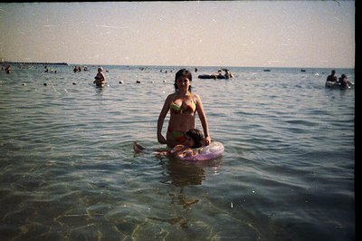 A woman in a patterned bikini cradles a child on an inflatable ring in shallow, turquoise water. Background shows a bustling ...