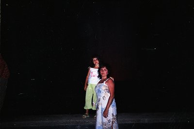 A young girl and woman stand on a concrete surface, illuminated by an unseen light source. The girl wears a yellow t-shirt an...