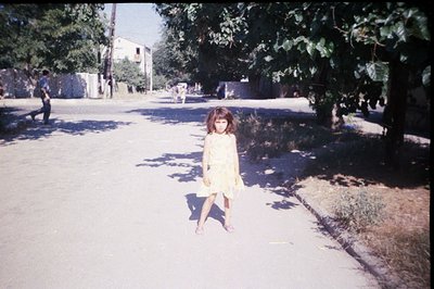 A young girl in a pale yellow dress stands on a cracked asphalt road. Shadows from trees dapple the scene. Two boys are visib...