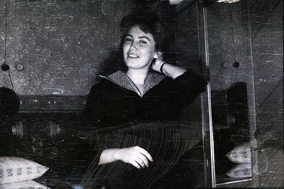 A young woman seated near a window, appearing on a train. She wears a dark, collared dress. The photograph exhibits age with ...