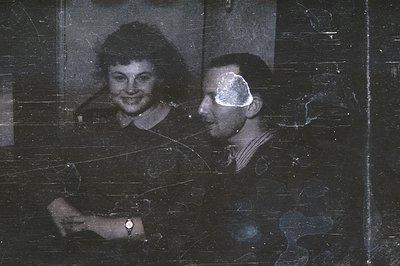 A posed portrait of a young couple, likely taken in the 1940s. The woman wears a dark dress and a wristwatch; the man is in a...