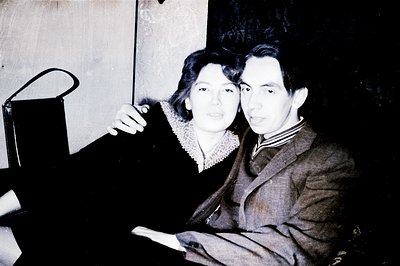 A posed portrait of a couple in what appears to be formal attire, circa 1950s. The woman wears a dark dress with lace detaili...