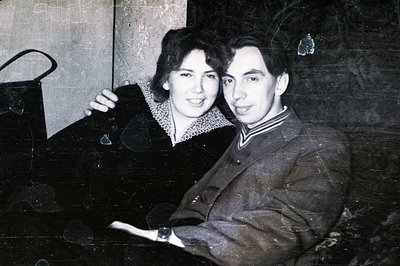 A formally dressed couple poses close together. The woman wears a dark dress with a ruffled collar, while the man is in a sui...