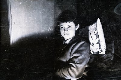 A young woman in a dark suit stands formally with arms crossed, posed near a door. Visible grain and scratches indicate age &...