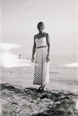A young woman stands on a beach, facing the camera with a slight smile. She wears a polka-dot sundress and sandals, posed aga...