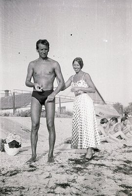 A man in swim trunks and a woman in a patterned, long dress stand on a sandy beach. A lifeguard station and sunbathers are vi...