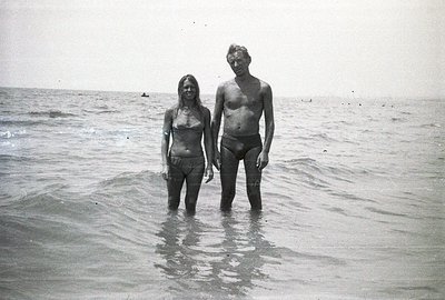 A couple stands chest-deep in shallow water, facing the camera. The man, shirtless, wears dark swim trunks; the woman, a stri...