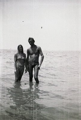 A man and woman wade in shallow water, likely at a seaside location. Both are wearing swimwear typical of the 1960s-70s. The ...