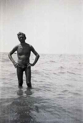 A man stands knee-deep in shallow water, looking directly at the camera. He wears a brief swimsuit, hands on his hips, posed ...