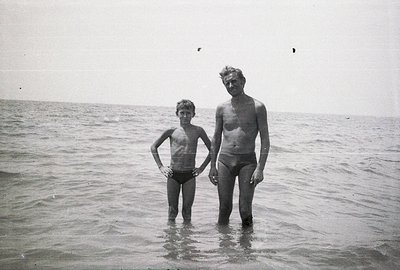 A boy and man stand waist-deep in shallow water, seemingly on a beach. Both are unclothed except for dark swim trunks. The sc...