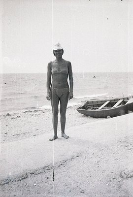 A young woman stands on a sandy beach, wearing a vintage swimsuit and a sailor’s cap. A small, wooden boat rests on the sand ...
