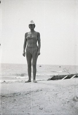 A woman stands on a beach, facing the viewer, wearing a modest, one-piece bathing suit and a headscarf. Gentle waves lap the ...