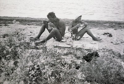 Two shirtless men sit on a patterned blanket during a picnic, enjoying a meal. A body of water stretches into the distance. L...