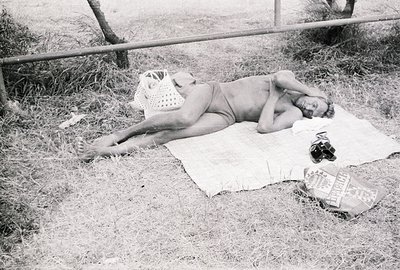 A man lies prone on a patterned blanket, seemingly resting outdoors. He's partially obscured by foliage and a woven bag. A ca...