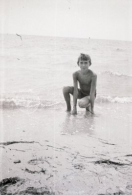 A young, shirtless boy kneels in shallow water, facing the camera. The composition is simple, focusing on the child and the i...