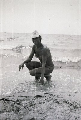 Woman in a vintage swimsuit and wide-brimmed hat kneels in shallow water at a sandy beach. The composition focuses on the fig...