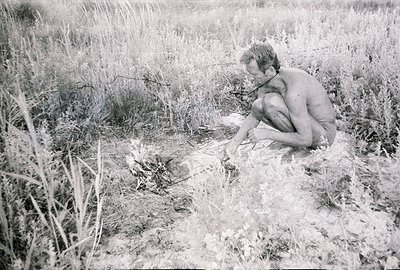 A shirtless man kneels amidst tall, overgrown grass and foliage, carefully tending to a small fire. He appears focused on the...