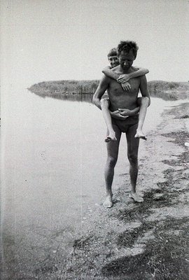 Man carries a young boy on his shoulders along a sandy beach. Simple, candid moment captured likely in the 1960s or 70s. Shal...
