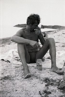 Young man seated on a sandy beach, appears to be consuming food. Wearing patterned swim trunks. A small jar sits nearby on a ...