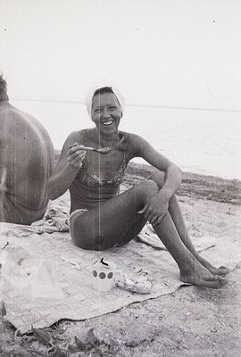 A smiling woman, seated on a beach blanket, enjoys a meal outdoors. She wears a vintage-style floral bikini and a headscarf. ...
