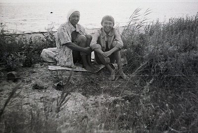 Two figures, an adult woman and a young boy, seated on a patterned blanket in a coastal landscape. The woman wears a printed ...