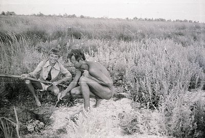 Two young men, one partially clothed, examine a small burrow or hole in a grassy field. The man in shorts holds a stick, whil...