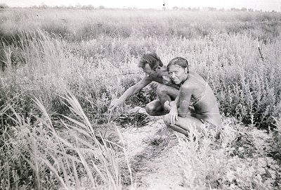 Two figures, a man and a woman, kneel among tall grasses and vegetation. The man examines the ground, while the woman gazes t...