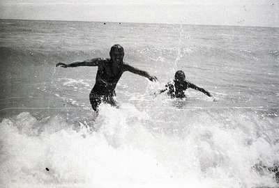 Two children are joyfully wading through surf, arms outstretched. The monochrome image captures a moment of playful energy ag...