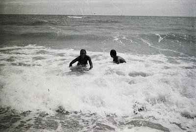 Two figures navigate churning ocean waves. The figures appear to be wading or swimming in rough water, likely enjoying a seas...