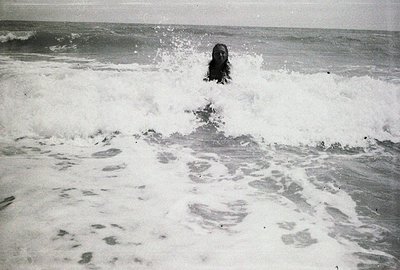 A figure emerges from crashing waves, mid-stride. The monochromatic image captures a moment of motion with turbulent surf and...