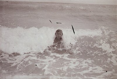 A young man stands in mid-chest-deep ocean water, partially submerged by a breaking wave. The image shows a vintage, grainy a...